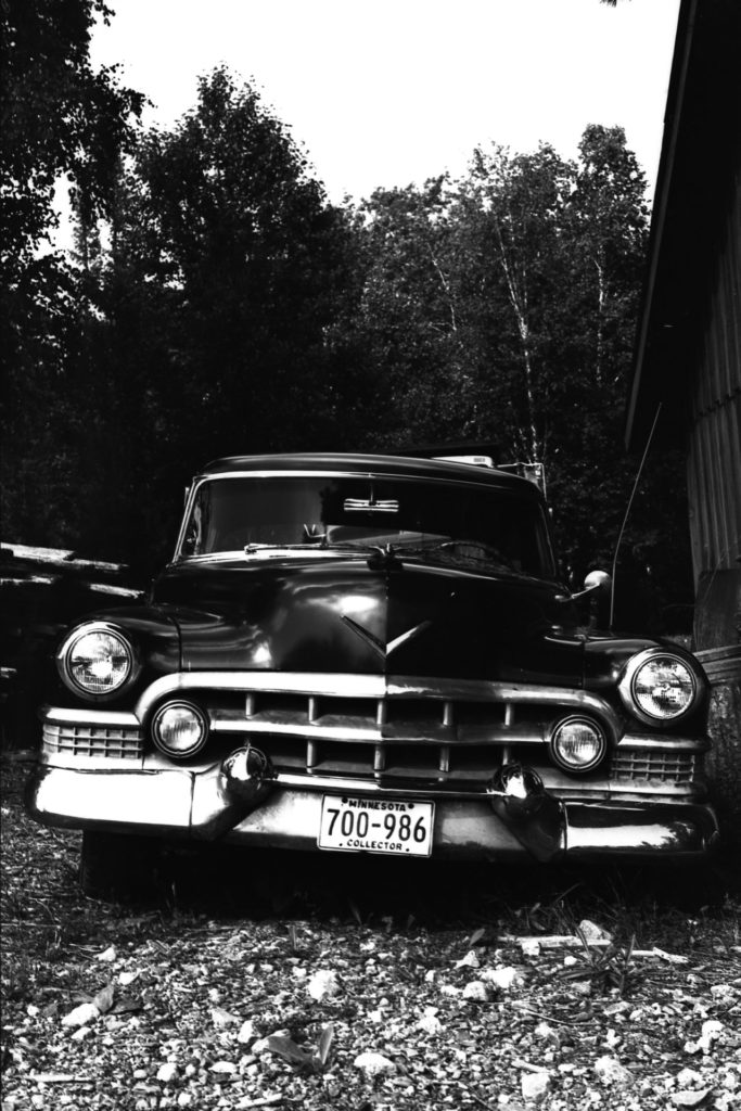 A black and white photo of an old American car parked in front of some tall trees.