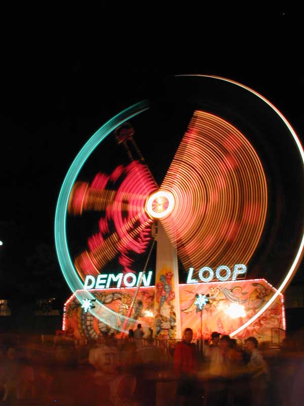 A color, long-exposure shot of a Midway ride called Demon Loop. The ride is going in a vertical circle. The left hand of the circle has long blue streaks of light. The right side of the circle has long yellow streaks of light.