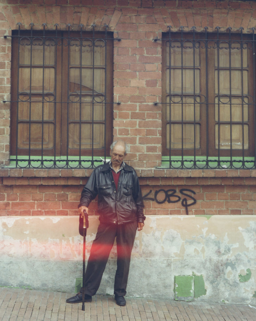A close up version of the previous photo. A color photo of a Bogotano man leaning on a cane standing in front of a brick building