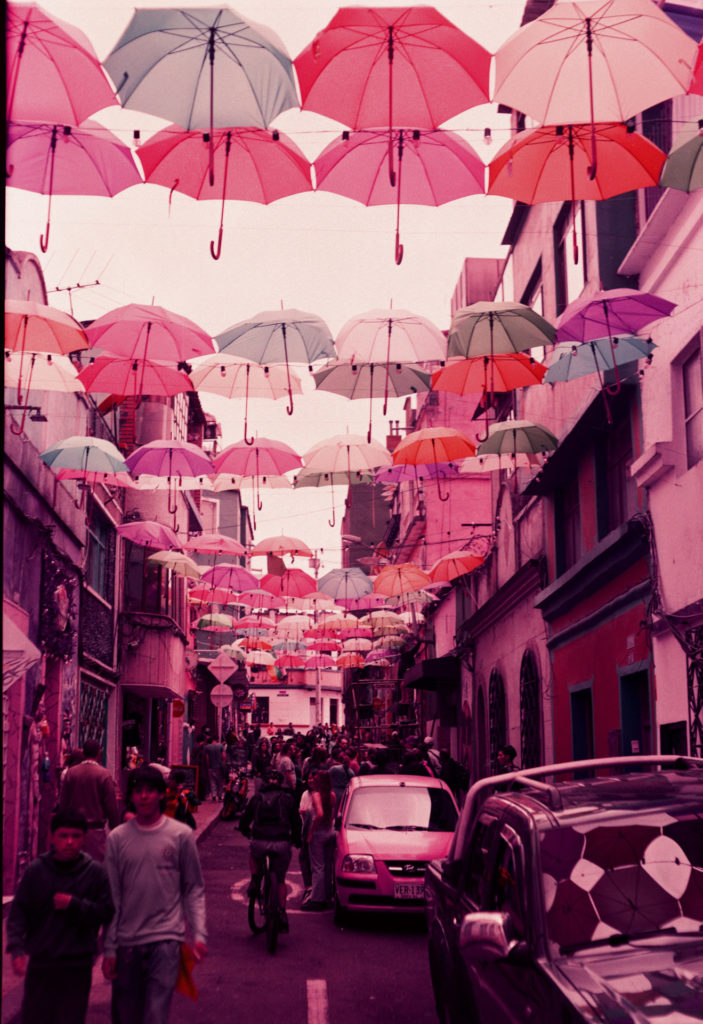 A color photo of a narrow street decorated with umbrellas. The color of the photo is tinted purple.