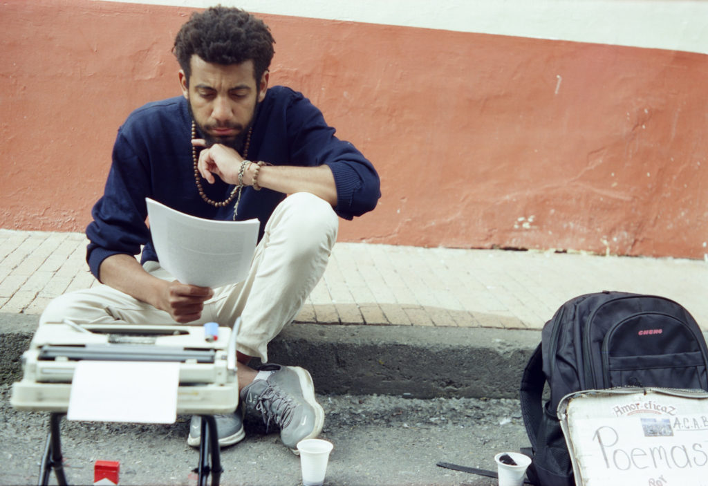 A young man sits on a street curb, reading a piece of paper. In front of him is a typewrite and a sign. The sign says that the man will write you a poem if you pay.