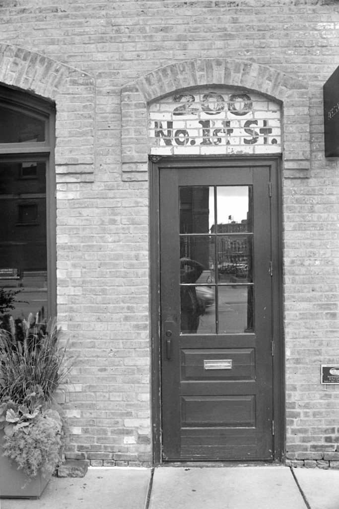 A black and white photo of a black wooden door set in a brick building in the North Loop area of Minneapolis. Above the door painted on bricks is the address, "200 No. 1st St."