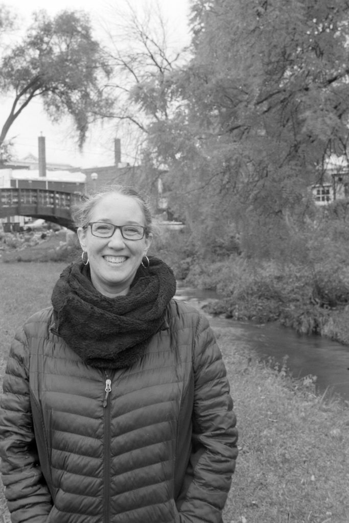 A black and white photo portrait of a person. Behind them is a creek, which is crossed by a bridge. The background is not very sharp, but the objects are recognizable.