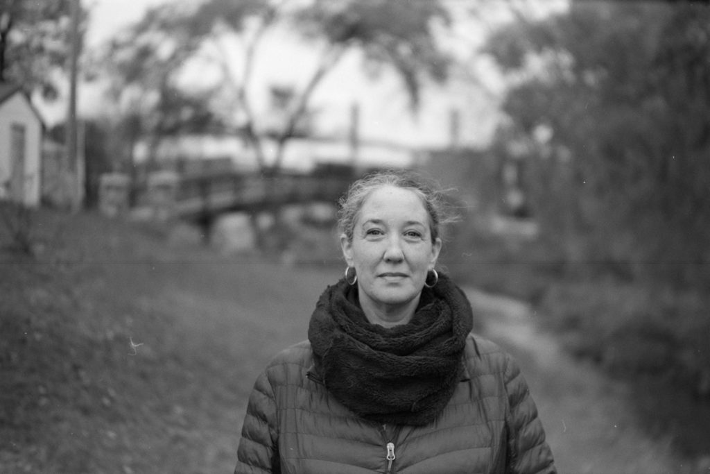 A black and white photo portrait of a person. Behind them is a creek, which is crossed by a bridge. The background is quite blurry and looks somewhat swirly.