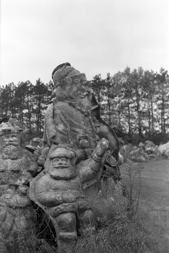 A black and white photo of a pile of Santa Claus fiberglass molds. They vary in size from 4 feet to 8 feet tall. Some are covered in lichen.