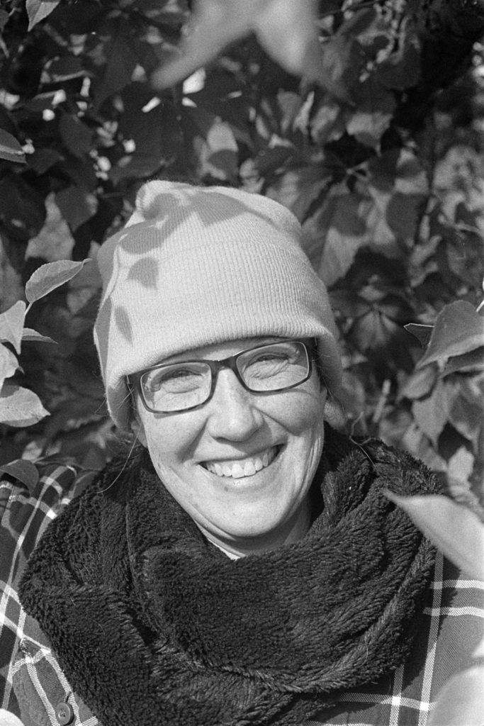 A close up black and white portrait of a person surrounded by the leaves on an apple tree. The person is wearing a scarf, a knit hat and glasses. They are smiling.
