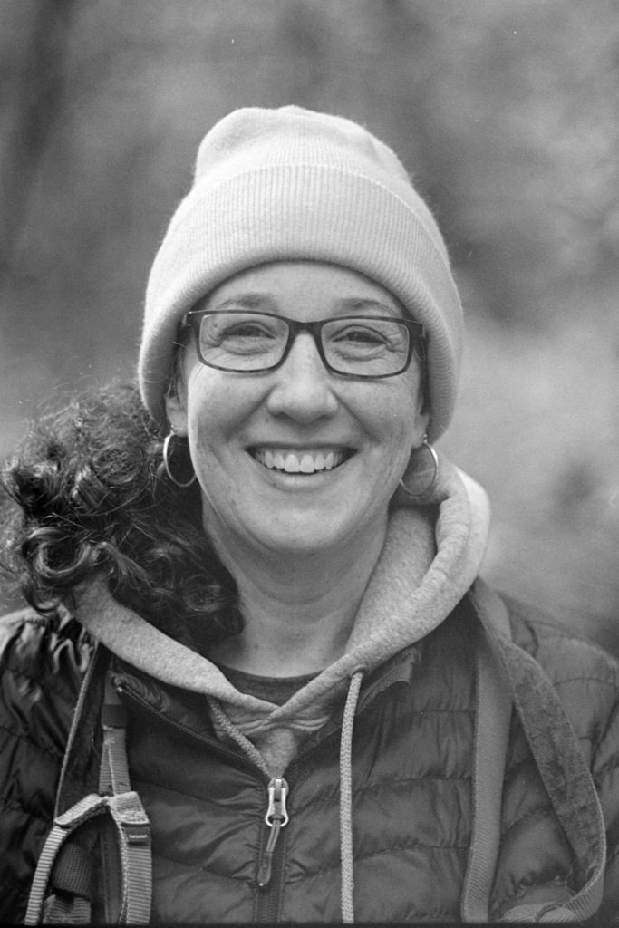 A black and white close up portrait photo of a person. They are wearing a grey sweatshirt, dark jacket, a knit hat and glasses. The background is entirely blurry.