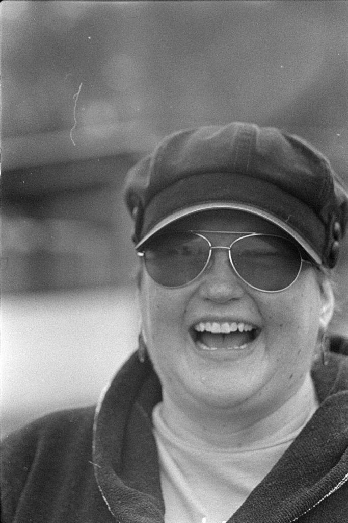 A black and white portrait photo of a person laughing. They are wearing a dark sweatshirt, dark sunglasses and dark hat. The person is in focus, but somewhat soft. The background is blurry.