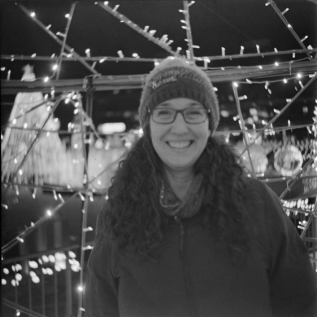A black and white photo of a woman at night. The woman is wearing a dark jacket, glasses and a knit hat. She is standing in front of a display of holiday lights. The entire photo is slightly out of focus.
