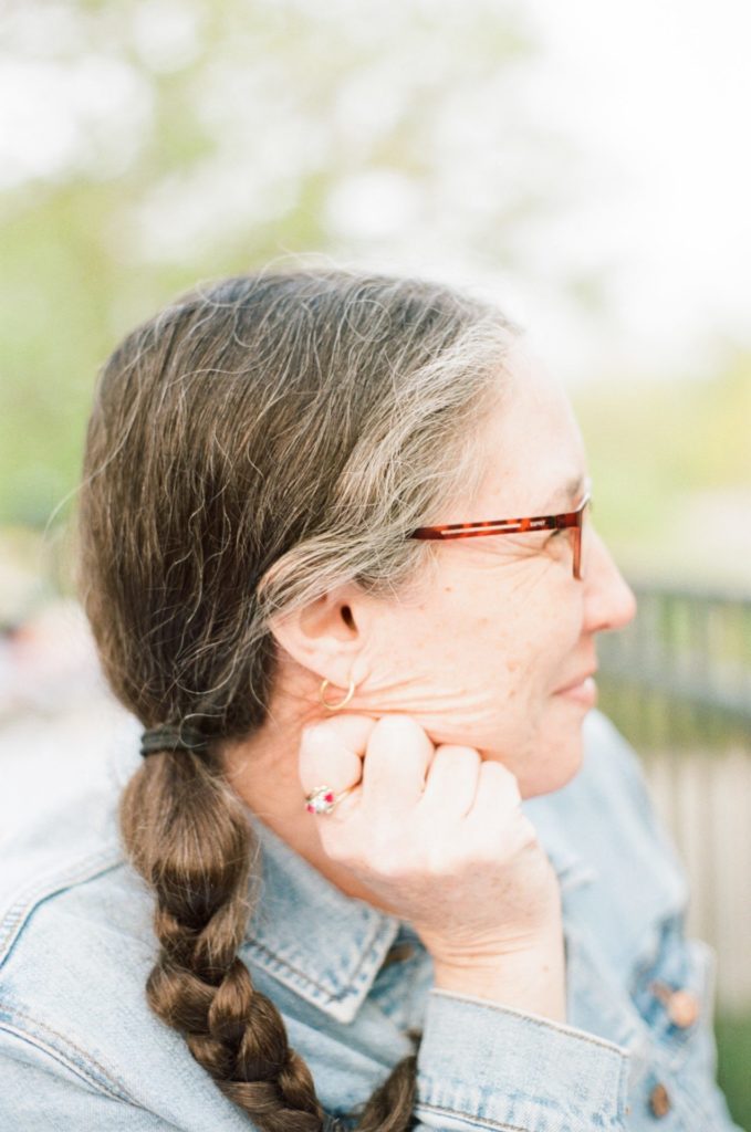 A color profile portrait of a woman. Her brown and grey hair is braided. She is wearing glasses and a faded jean jacket. The background is a blurry tree.