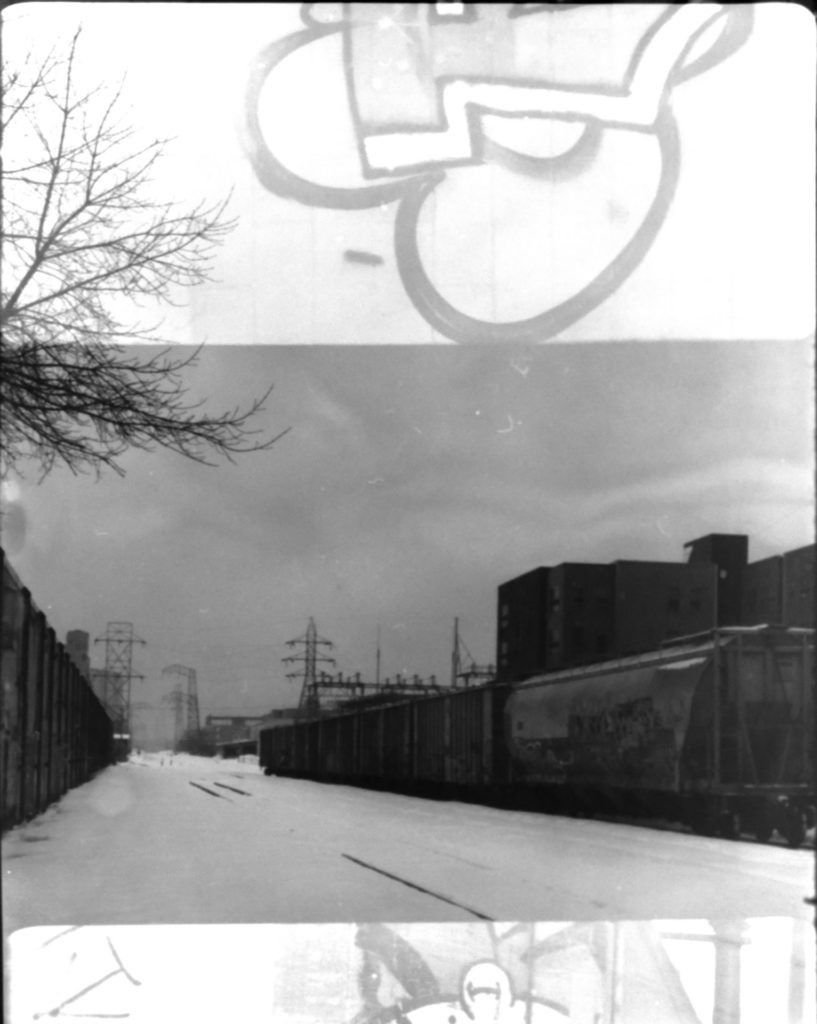 A black and white double exposure. Horizontally there's a wide shot of some grain-carrying train cars sitting in a rail yard. Horizontally across the frame is a close up of some graffitie on one of the cars.