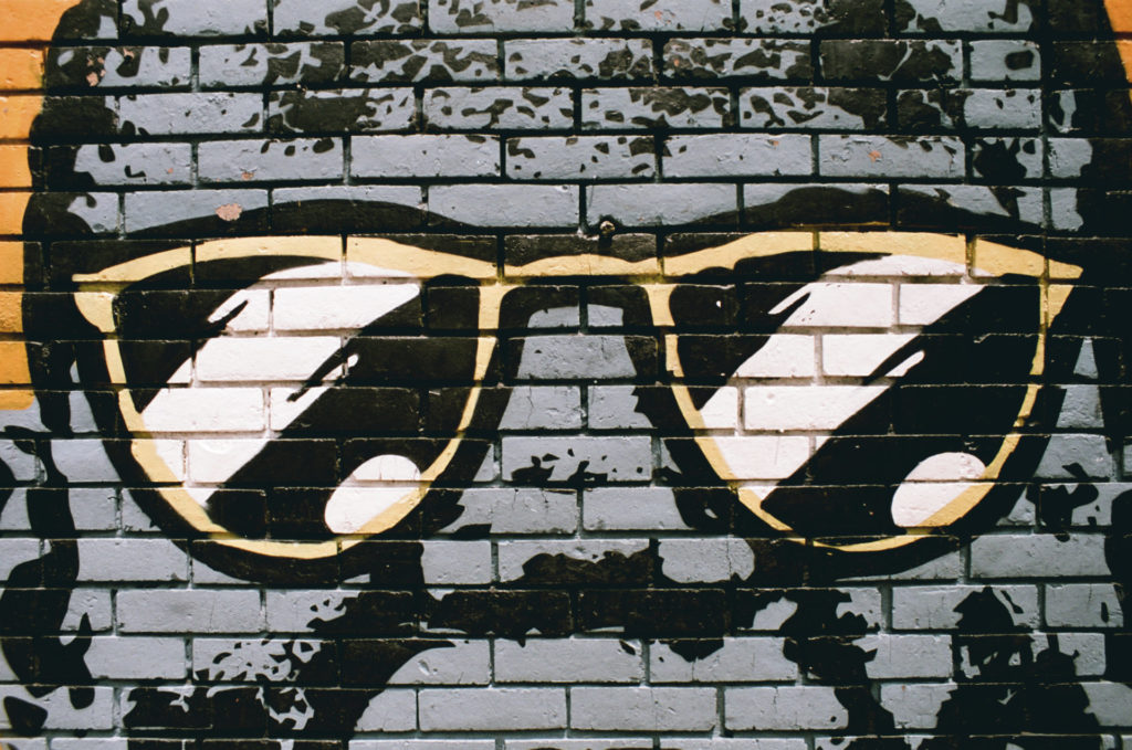 A color photo of a street art mural painted on bricks. The photo shows part of the paining, mostly gold-rimmed sunglasses.