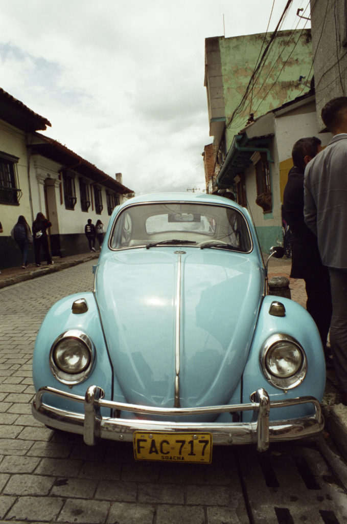 A color photo of a light blue classic VW Bug parked on a street in Bogotá, Colombia