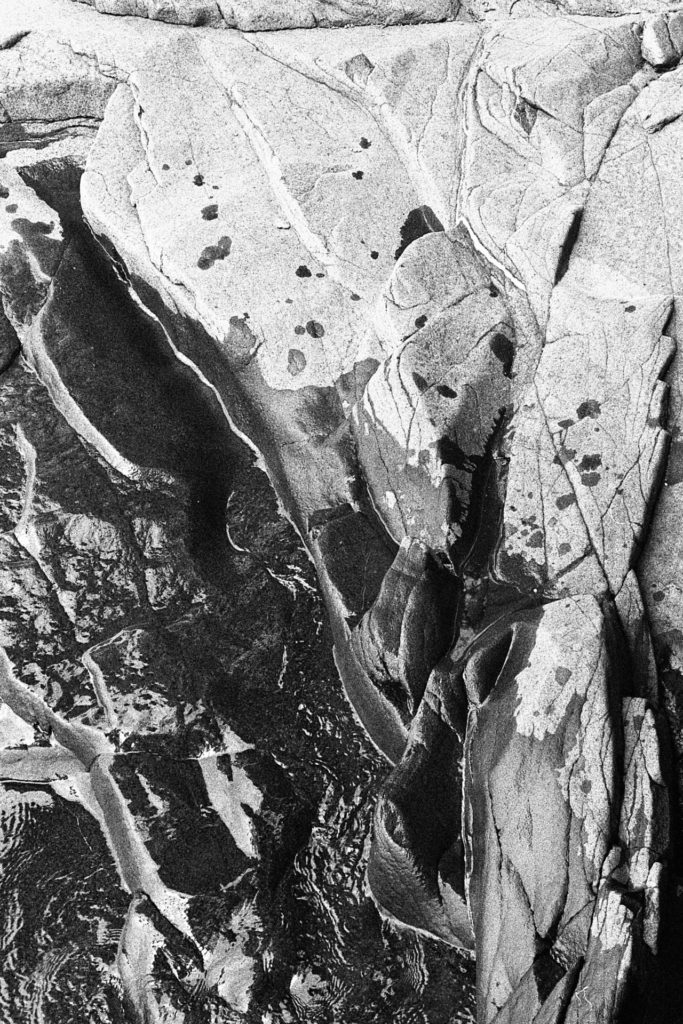 A black and white photo of the waters of Lake Suprior lapping the grey granite shore. Where the water has recently touched the rocks the rocks are black.