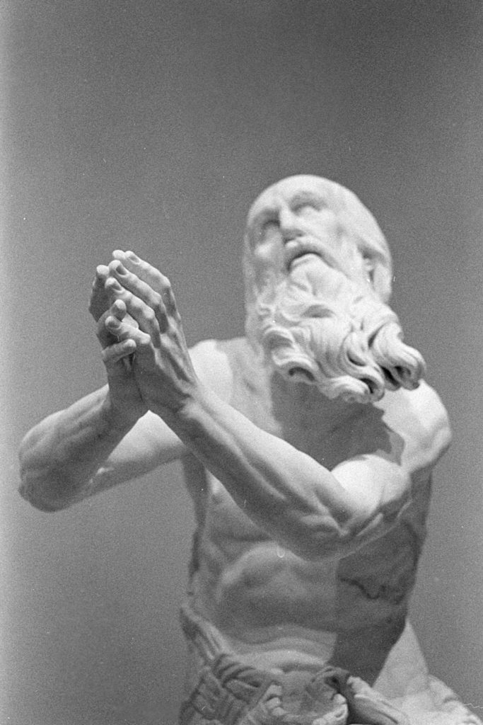 A black and white photo of a white marble statue of a bearded man. His hands are held out in front of him, arms bent, as if in prayer. The hands are in focus but the head is slightly blurry.