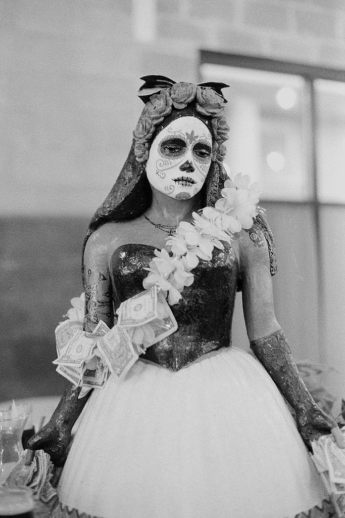 A black and white photo of a statue of a woman. She wears a shawl over her head covered in flowers. Her face is painted as if it's a skull -- in Dia de los Muertos style -- with white skin and black eye sockets. She wears a black corset and a white lace skirt. Crossing her body is a bandoleier of flowers and money. She holds dollar wads of dollar bills in both hands.