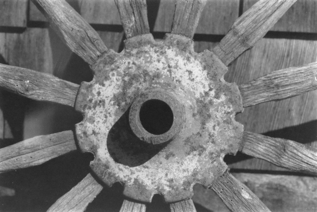 A landscape black and white picture of a wooden wagon wheel. The metal hub is in the center of the photo and the wooden spokes radiate outwards. The rim of the wheel is not visible. In this scan of the negative the details of the wood grain are blurry and the contrast of the picture is low.