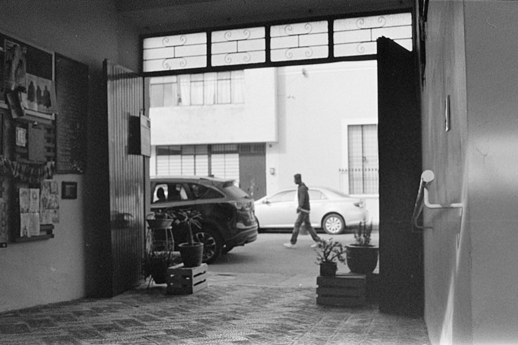 A black and white photo of a Guadalajara street seen from the inside of a cafe courtyard. Two large wooden doors are open and surrounded by potted plants. A man is walking down the street visible through the doors.