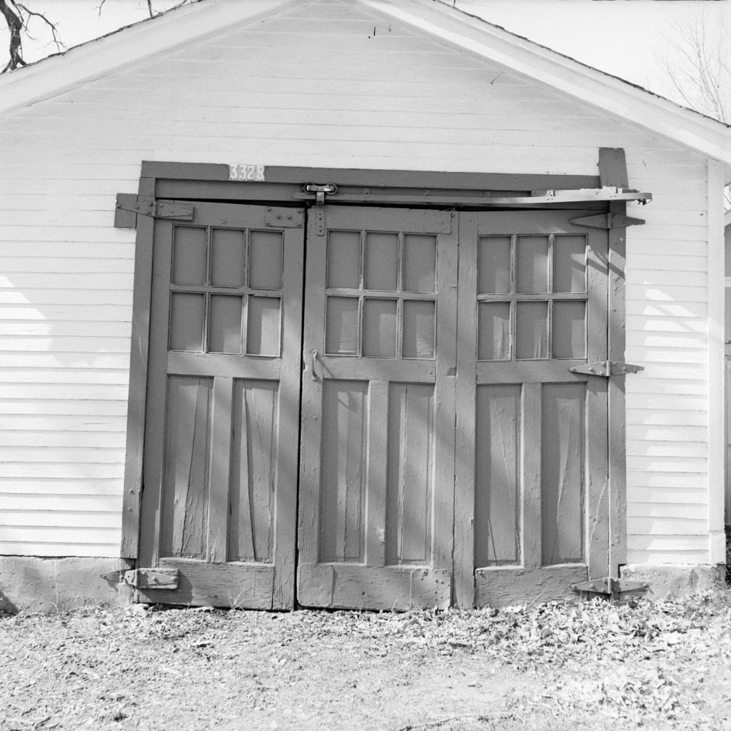 A black and white photo of an old garage door comprised of 3 small wooden doors on a rolling track.