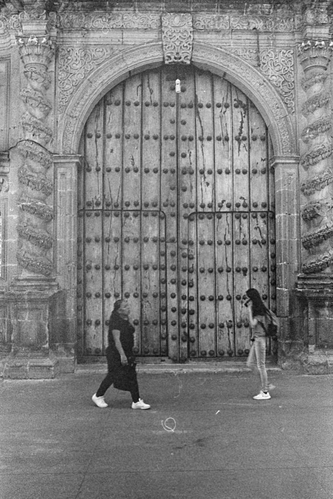 A black and white photo of a large wooden door to a church. The door is closed. Two women walk in opposing directions in front of the door. The photo is very grainy.