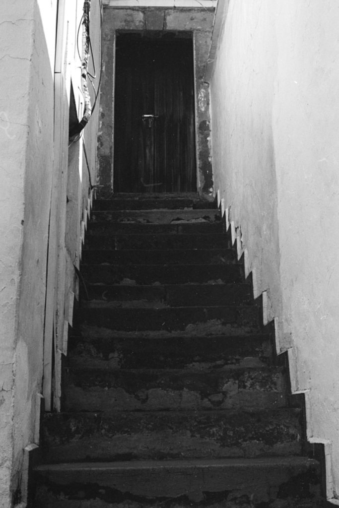 A dark stone staircase flanked by two white walls leads to a closed and bolted wooden door.