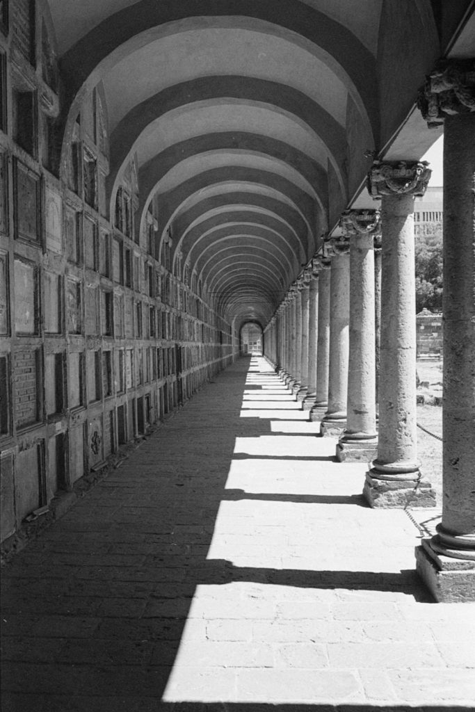 A black and white photo of a long outdoor walkway with an arched roof. On the left, in shadow, are lines of tombs along the wall. On the right, in light, are pillars.