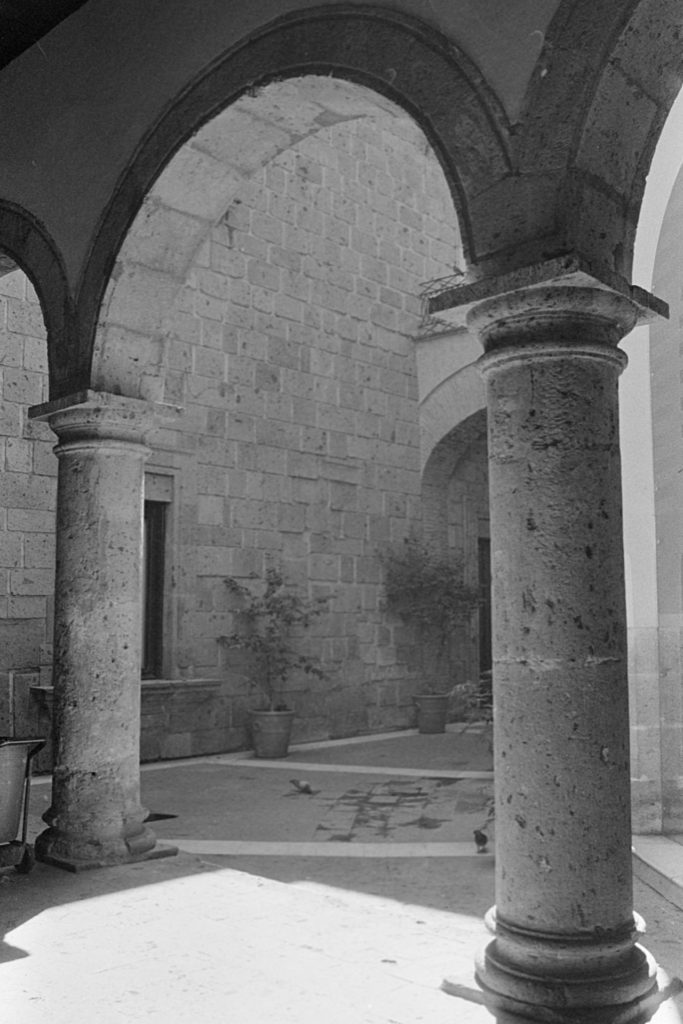 A black and white photo of a stone courtyard, mostly in shade. Some strong sunlight makes a bright patch between two pillars.