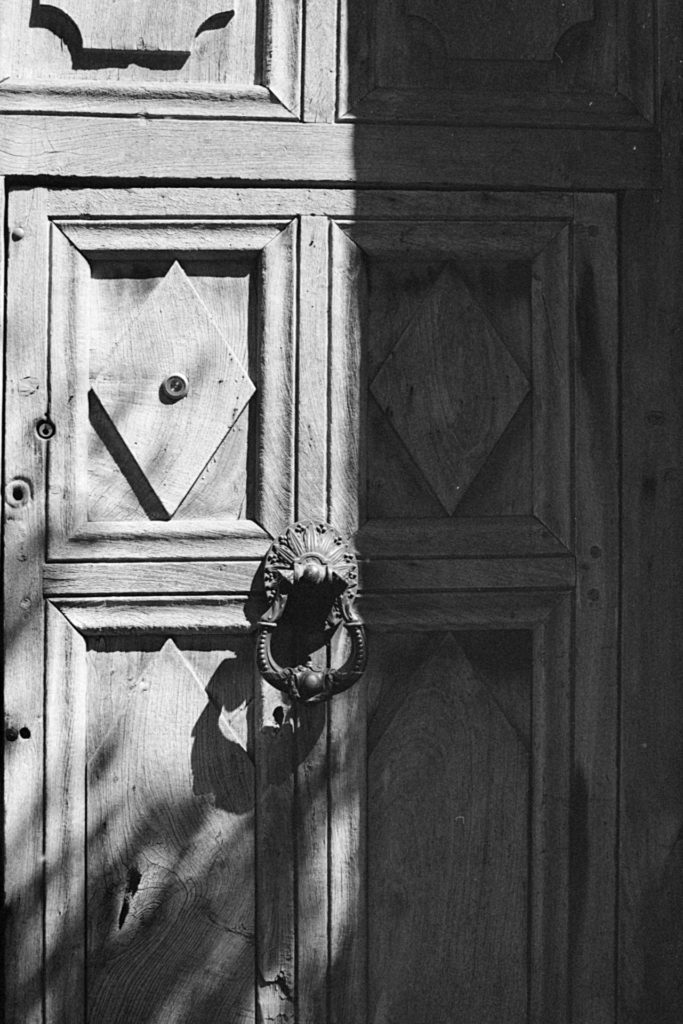 A black and white photo of an old, weathered wooden door. It is half in shadow. In the center is a large metal knocker.