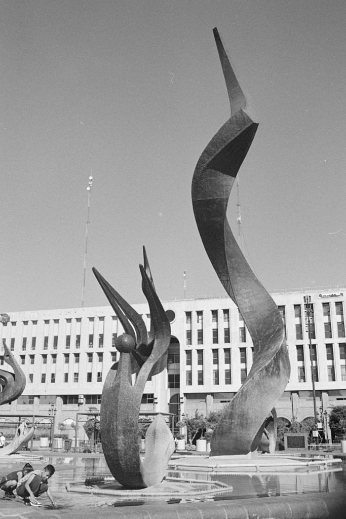 A black and white photo of a large outdoor sculpture surrounded by water. Two children are playing in the water near a smaller sculpture.