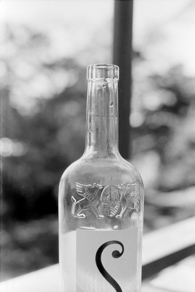 A black and white photo of a clear glass wine bottle with small drops of wine in the neck.