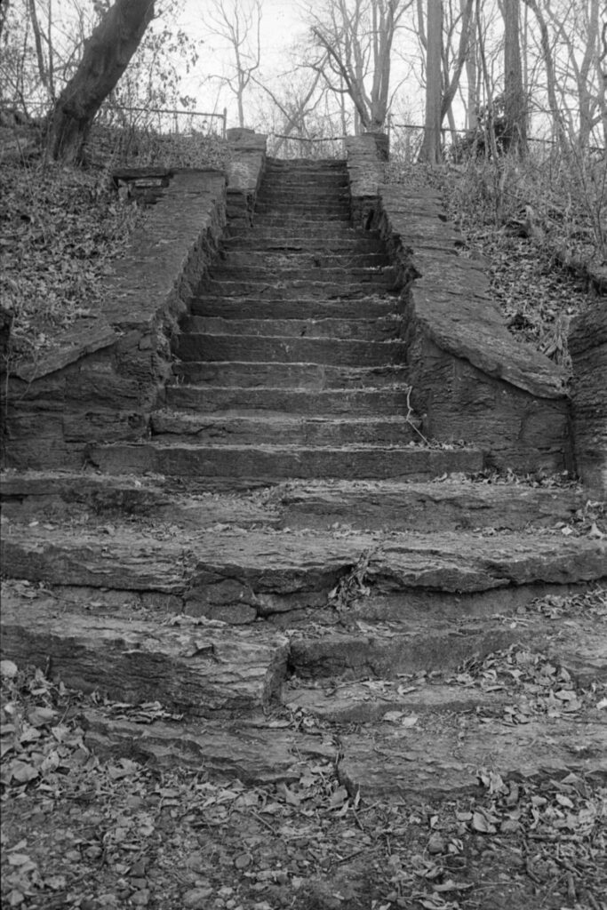 A black and white photo of a weathered stone staircase set into a hill. Though usable, some of the steps are crumbling away due to erosion.